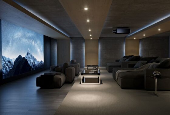 A large home theater with a large movie screen, a Sony projector, and large gray chaise lounges for seating.
