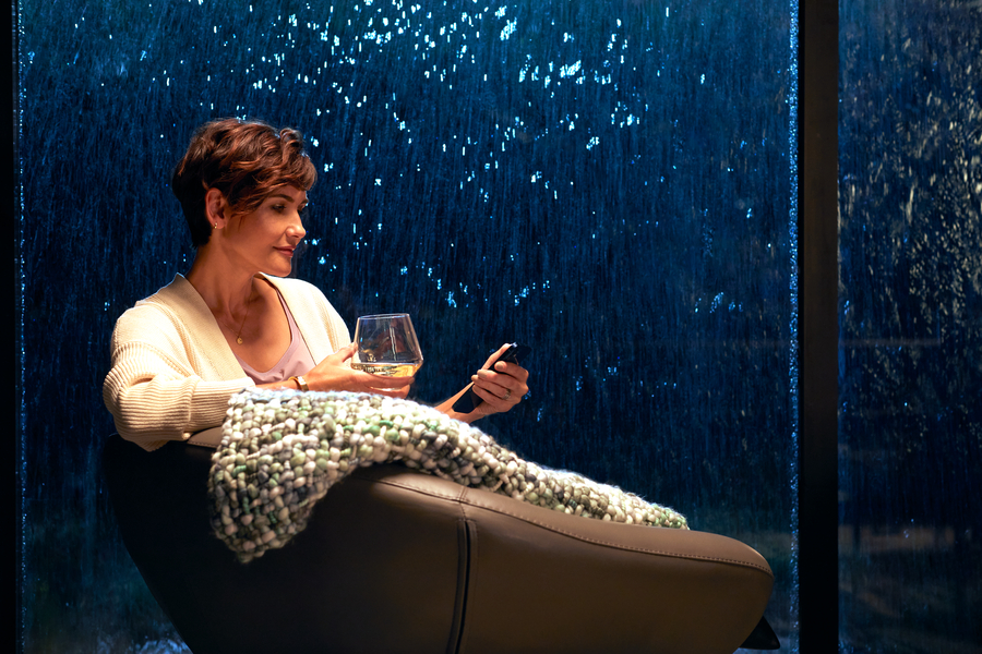 A woman sitting in a chair next to a window with rain pouring down. She’s holding a drink and the Savant remote.